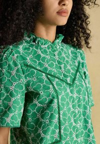 Blusa verde ricamata con un motivo floreale, dettagli in pizzo testurizzato e colletto a scallop. Maniche corte e design morbido.