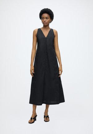 Femme debout portant une robe midi noire sans manches à décolleté en V et des sandales noires à talons sur un fond uni clair.