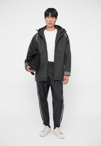 Svart jacka med ljusgrå detaljer, två framfickor, bärs över en vit skjorta. Svarta, glänsande träningsbyxor med vita sidostripes och krämfärgade sneakers.