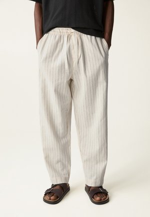 Osoba ubrana w luźne, kremowe spodnie w cienkie pionowe paski, czarną bluzkę i ciemnobrązowe sandały na klamrę, stojąca z rękami w kieszeniach.