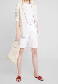 Blazer en lin beige, t-shirt blanc, shorts blancs à revers, sandales rouges à lanières et un sac en crochet avec une poignée en bambou.