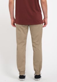 Pantalons beige fuselés en matériau léger avec des poignets élastiques ; portés avec une chemise bordeaux et des baskets noires.
