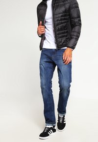 Svart quiltad jacka, vit t-shirt, blå denimjeans och svarta sneakers med vita ränder. Avslappnad outfit med kroppsnära design och upprullade ärmar.