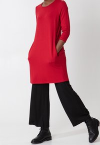 Rode tuniekjurk met driekwartmouwen, zijzakken en een ontspannen pasvorm, gecombineerd met brede zwarte broek en zwarte veterschoenen.