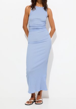 Robe maxi bleu clair côtelée avec une coupe ajustée et des détails froncés sur le côté, associée à des tongs blanches sur un fond uni.