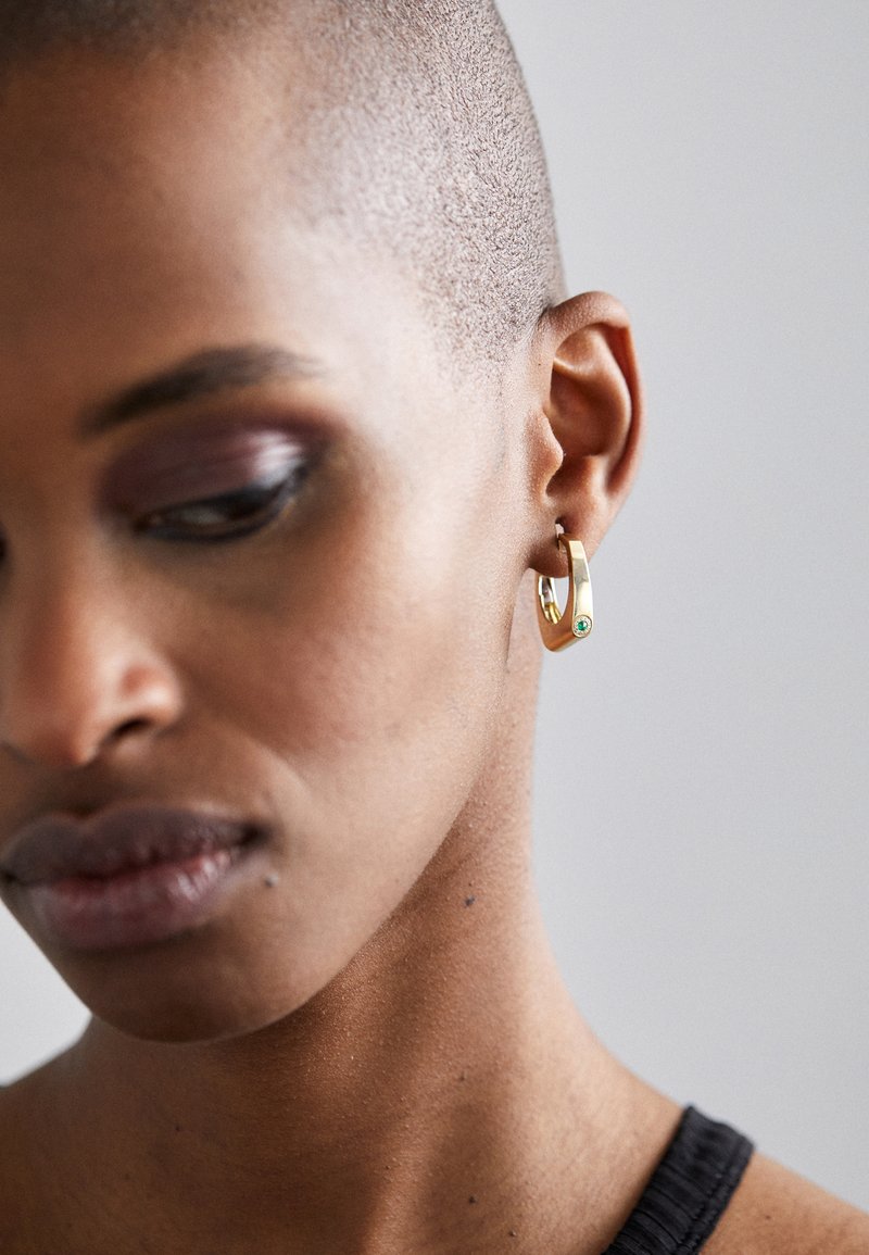 Cercei hoop aurii cu o formă geometrică și un mic accent de piatră verde, purtați pe o fotografie în prim-plan a urechii unui model.