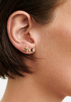 Korva, jossa kaksi kultista nappikorvakorua, jotka on muotoiltu tähdeksi ja sirpiksi, kumpikin koristeltu pienillä kirkkailla jalokivillä.