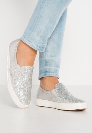 Fødder iført lyseblå jeans og sølvglitrende slip-on sneakers med hvide såler mod en ensfarvet lys baggrund.