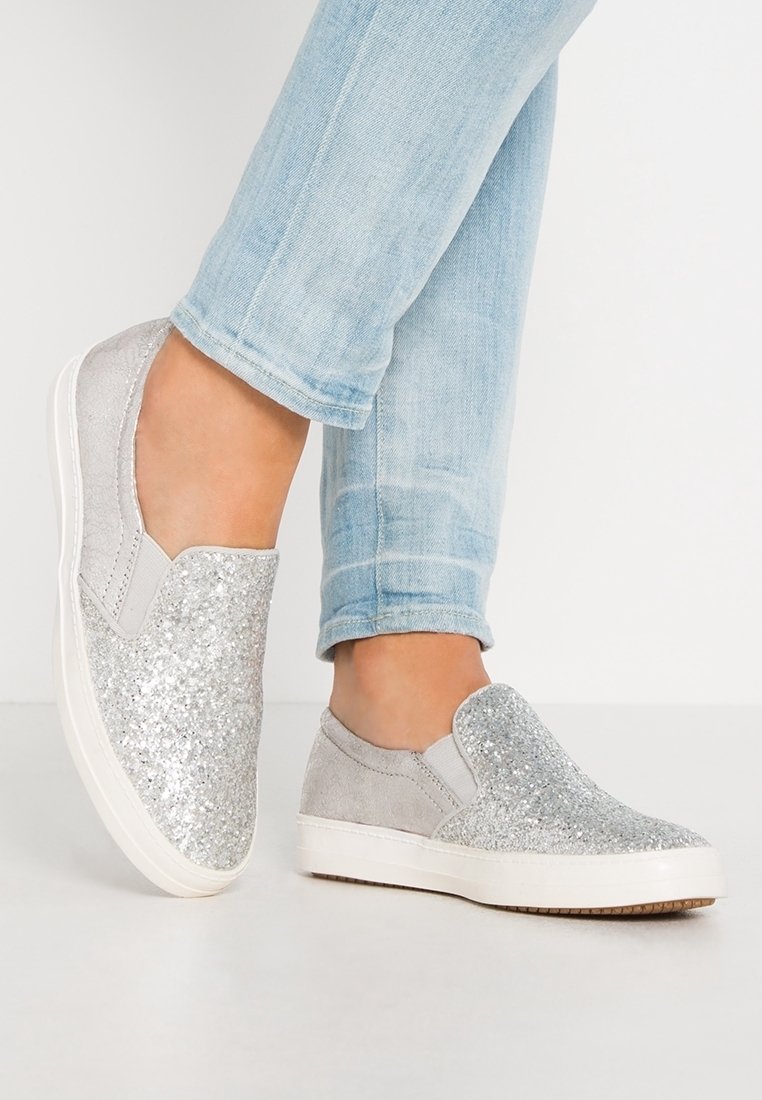 Pieds portant un jean bleu clair et des baskets slip-on argentées pailletées avec des semelles blanches sur un fond clair uni.