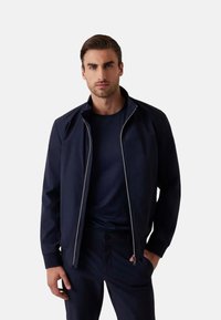 Giacca zip-up blu navy con alto colletto e sottili accenti bianchi. Realizzata in tessuto liscio, presenta cuciture orizzontali e una vestibilità slim.