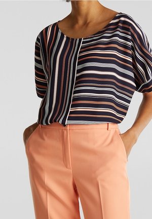 Mujer que lleva una blusa de manga corta a rayas en azul marino, blanco y naranja, metida dentro de unos pantalones de talle alto color melocotón, con las manos en los bolsillos.
