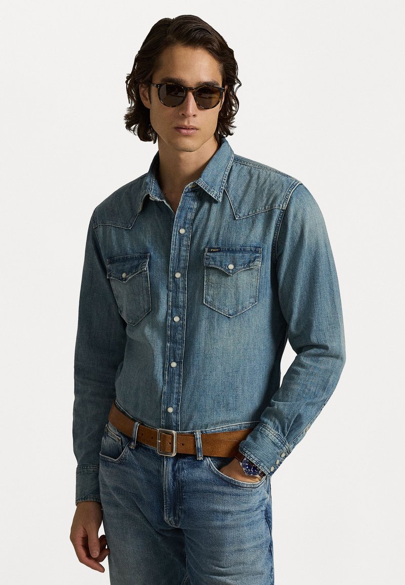 Heledsinine denim särk pikkade varrukatega, nööbiga ja kahe rinnataskuga. Seljas pruun nahkvöö ja heledate denim teksadega.