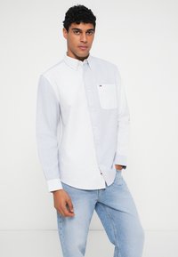 Bărbat cu păr negru, creț, îmbrăcat într-o cămașă cu dungi albastre și albe, cu nasturi, și blugi albaștri deschiși, stând în fața unui fundal alb simplu.