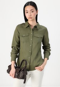 Camicia verde oliva con bottoni, due tasche sul petto, maniche lunghe e bottoni bianchi, abbinata a una borsa di pelle marrone scuro.