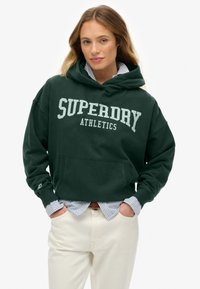 Mujer con pelo largo vistiendo una sudadera de atletismo Superdry de color verde oscuro sobre una camisa a rayas azul y pantalones de color blanco roto, con las manos en los bolsillos.