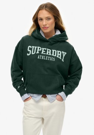 Woman with long hair wearing a dark green Superdry Athletics hoodie over a blue striped shirt and off-white pants, hands in pockets.