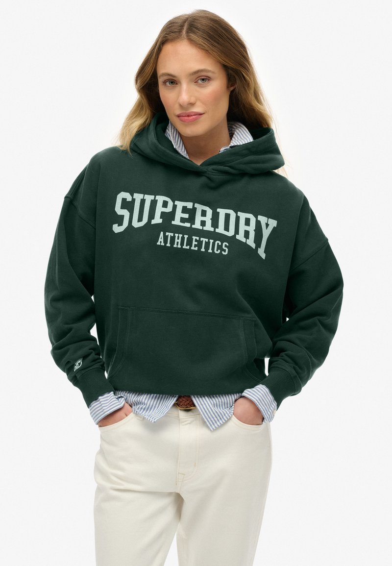 Mujer con pelo largo vistiendo una sudadera de atletismo Superdry de color verde oscuro sobre una camisa a rayas azul y pantalones de color blanco roto, con las manos en los bolsillos.