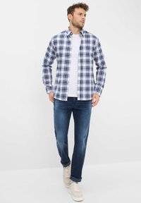 Camicia a quadri da uomo in blu, bianco e navy. Abbinata a jeans blu scuro e sneakers beige chiaro, con una maglietta bianca semplice sotto.