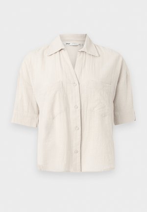 Világos bézs, rövid ujjú, gombos ing két mellzsebbel és hegyes gallérral, egyszerű háttér előtt bemutatva.