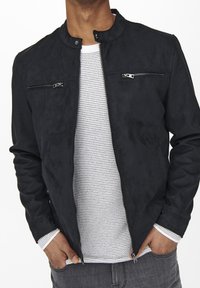 Giacca in suede nera con colletto alzato, due tasche sul petto con cerniera e una texture liscia, indossata sopra una camicia bianca con texture.