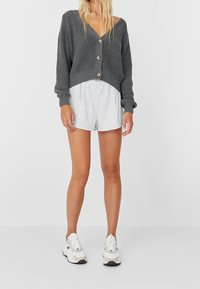 Grijze gebreide cardigan met diepe v-hals, drie knopen en geribde mouwen, gecombineerd met lichtgrijze short en witte sneakers.