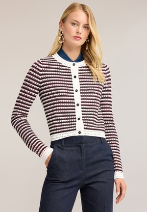 Blonde Frau mit goldenen Ohrringen, einem gestreiften, geknöpften Cardigan in Weiß, Schwarz und Dunkelrot und dunkelblauer Hose, eine Hand in der Tasche.