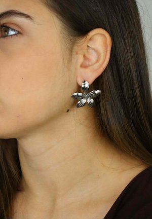 Orecchini d'argento a forma di fiore con petali testurizzati e un motivo centrale, indossati sull'orecchio di una persona con capelli castani lunghi.