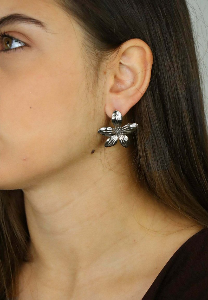 Srebrne kolczyki w kształcie kwiatów z teksturowanymi płatkami i centralnym wzorem, noszone na uchu osoby o długich brązowych włosach.