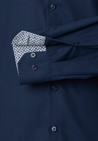 Marineblauw overhemd met opgestroopte mouwen om de binnenkant van de manchet te tonen, bedekt met een wit, blauw en zwart geometrisch patroon.