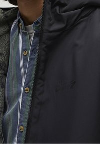 Czarna kurtka zapinana na zamek, nałożona na zieloną i niebieską koszulę w paski, z beżowymi guzikami i teksturowanym fleece'em w środku. Na kurtce nadrukowane logo.