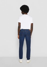 Vaqueros oscuros entallados, con bolsillos traseros, costuras de contraste y una etiqueta de cuero en la cintura. Combinados con una camiseta blanca.