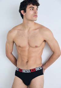 Caleçons noirs avec une ceinture blanche et rouge portant le logo "DIESEL". Tissu doux, coupe ajustée. Le modèle a les cheveux courts et bouclés, posé devant un fond neutre.