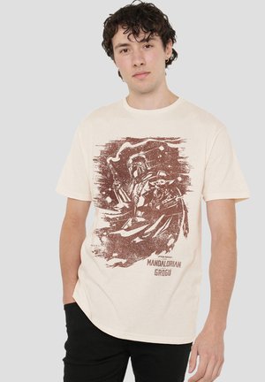 Junger Mann trägt ein cremefarbenes T-Shirt mit einer braunen Grafik von The Mandalorian, der Grogu aus Star Wars hält.