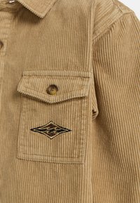 Giacca in velluto a coste beige con righe verticali, dotata di una tasca sul petto e un dettaglio del logo cucito in tonalità di marrone e nero.