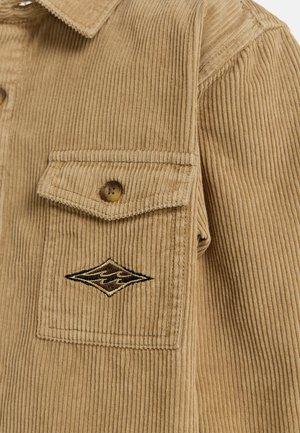 Corduroy jacket in beige with vertical ribbing, featuring a single chest pocket and a sewn logo detail in shades of brown and black.