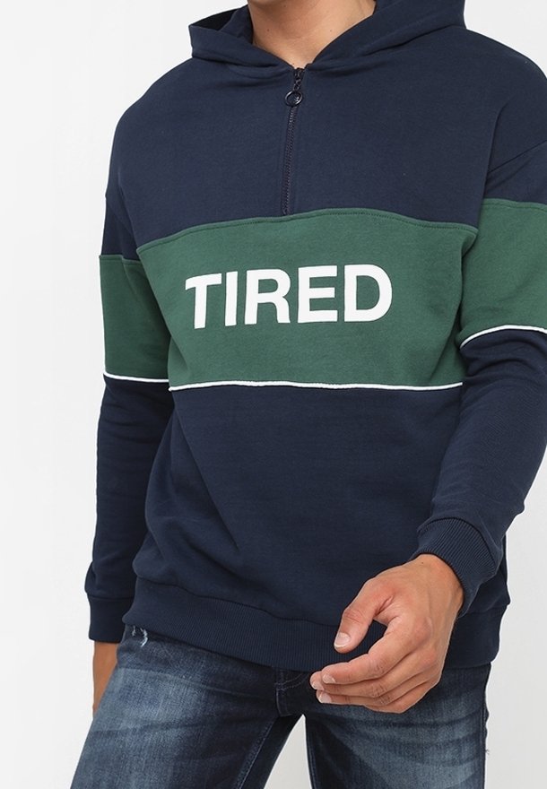 Homme portant un sweat à capuche bleu marine et vert avec fermeture éclair devant, texte blanc « TIRED » sur la poitrine, assorti à un jean bleu.