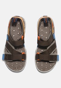 Sandalias deportivas marrones con suelas de textura hexagonal, correas ajustables marrones, acentos naranjas y etiqueta de marca azul. Diseño ligero.