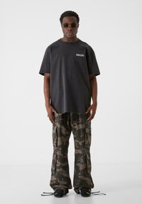 Sort oversized T-shirt med hvidt logo, parret med camouflage cargo bukser med flere lommer og en afslappet pasform. Sorte sko inkluderet.
