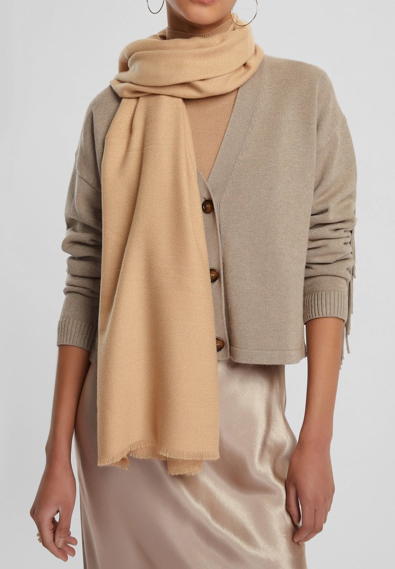 Cardigan in misto lana beige con scollo a V e bottoni, abbinato a una morbida sciarpa in tonalità sabbia drappeggiata, su una gonna di raso chiara con una leggera lucentezza.