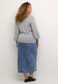 Grijze langemouwen top met ribgebreide accenten, gecombineerd met een blauwe denim maxi-jurk met achterzakken en een rechte silhoeët. Zwarte platte schoenen.