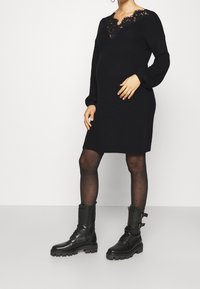 Czarna dzianinowa sukienka z koronkowym dekoltem, długimi rękawami i luźnym krojem, zestawiona z czarnymi, wysokimi do kolan, sznurowanymi botkami oraz rajstopami w kropki.