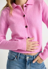 Rosa ullcardigan med krage, knappstängning, ribbade muddar och mjuk textur, i kombination med blå denimjeans.