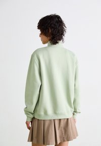 Sweatshirt vert clair en tissu doux, avec un col montant et une coupe décontractée, associé à une jupe plissée marron.