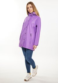 Giacca impermeabile viola con cappuccio, bottoni a pressione, due tasche laterali e un design leggero, indossata sopra jeans blu e sneakers.