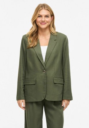 Lachende vrouw met lang blond haar, gekleed in een olijfgroene colbert en bijpassende broek over een wit topje, staand voor een effen achtergrond.