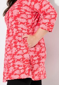 Czerwona tunika z różowym kwiatowym wzorem, z rękawami o długości trzech kwart i bocznymi kieszeniami. Miękka tkanina o swobodnym kroju i zaokrąglonym dole.