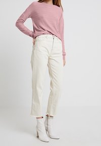 Maglione rosa testurizzato con maniche lunghe, abbinato a jeans bianchi a vita alta con orli sfrangiati e stivaletti bianchi alla caviglia.
