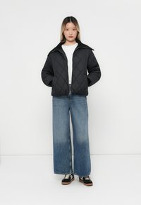 Giacca nera trapuntata con colletto, indossata sopra una camicia bianca. Abbinata a jeans blu a gamba larga e sneakers nere. Design minimalista, texture liscia.