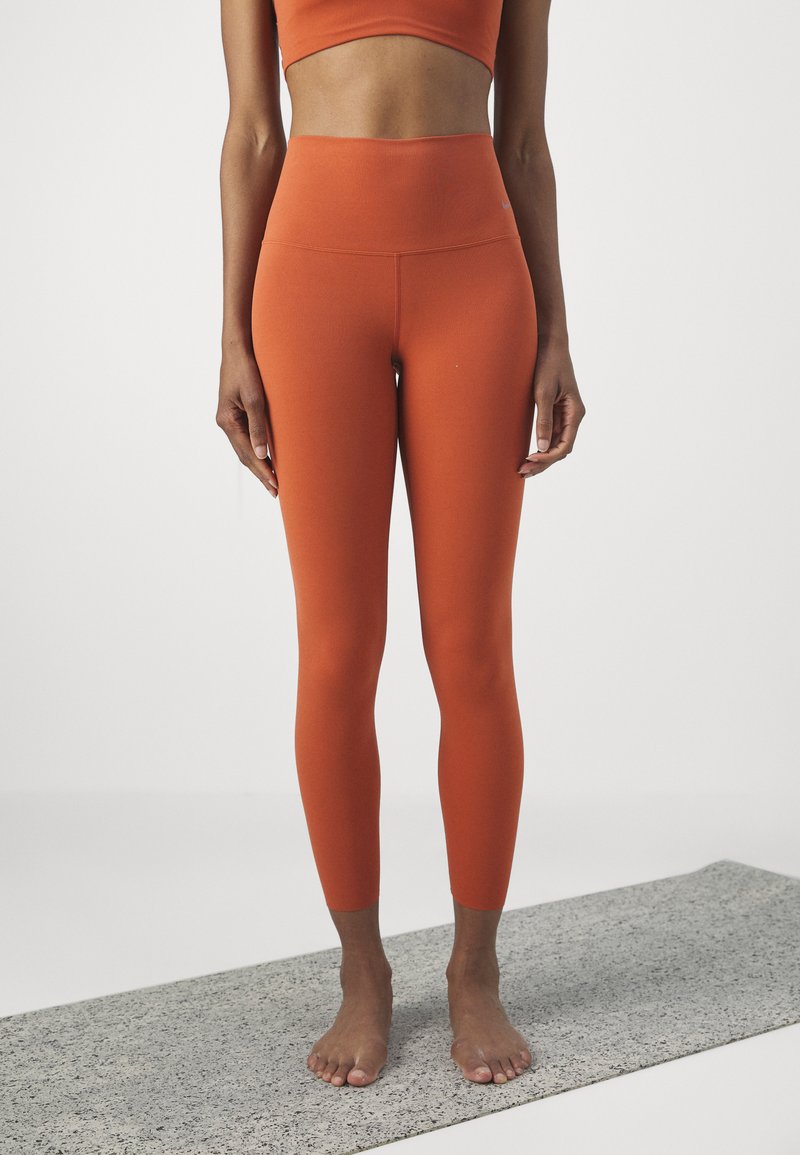 Leggings arancioni a vita alta con una texture liscia e un design aderente. Abbinati a un top cropped coordinato, esposti su un tappetino da yoga grigio.