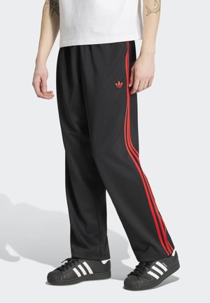 Czarne spodnie Adidas z czerwonymi paskami, noszone z czarnymi butami sportowymi Adidas z białymi paskami oraz białą koszulą widoczną w pasie.